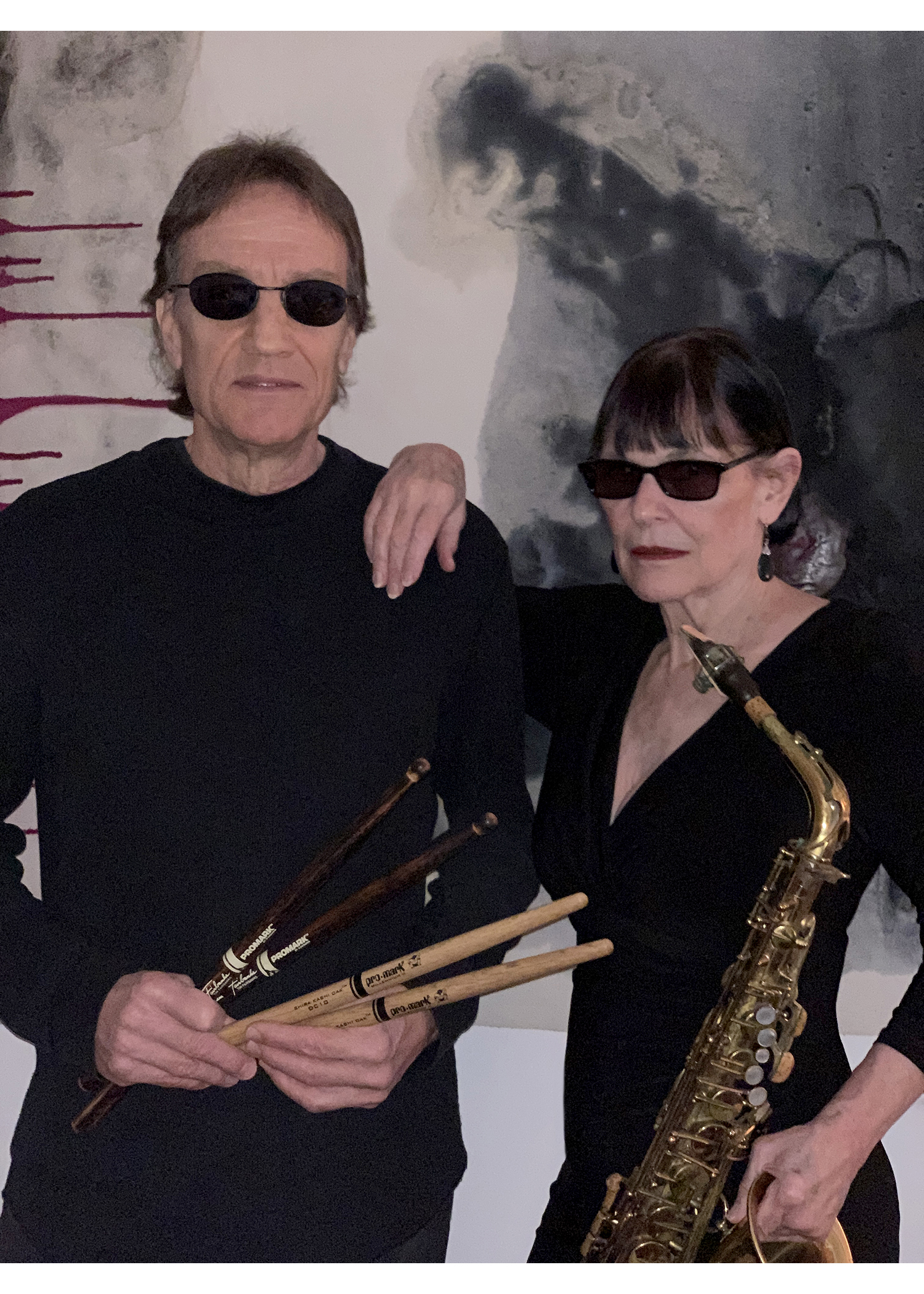 Two people in all black and sunglasses. The person to the left is holding drumsticks, and the person to the right is holding a saxophone.