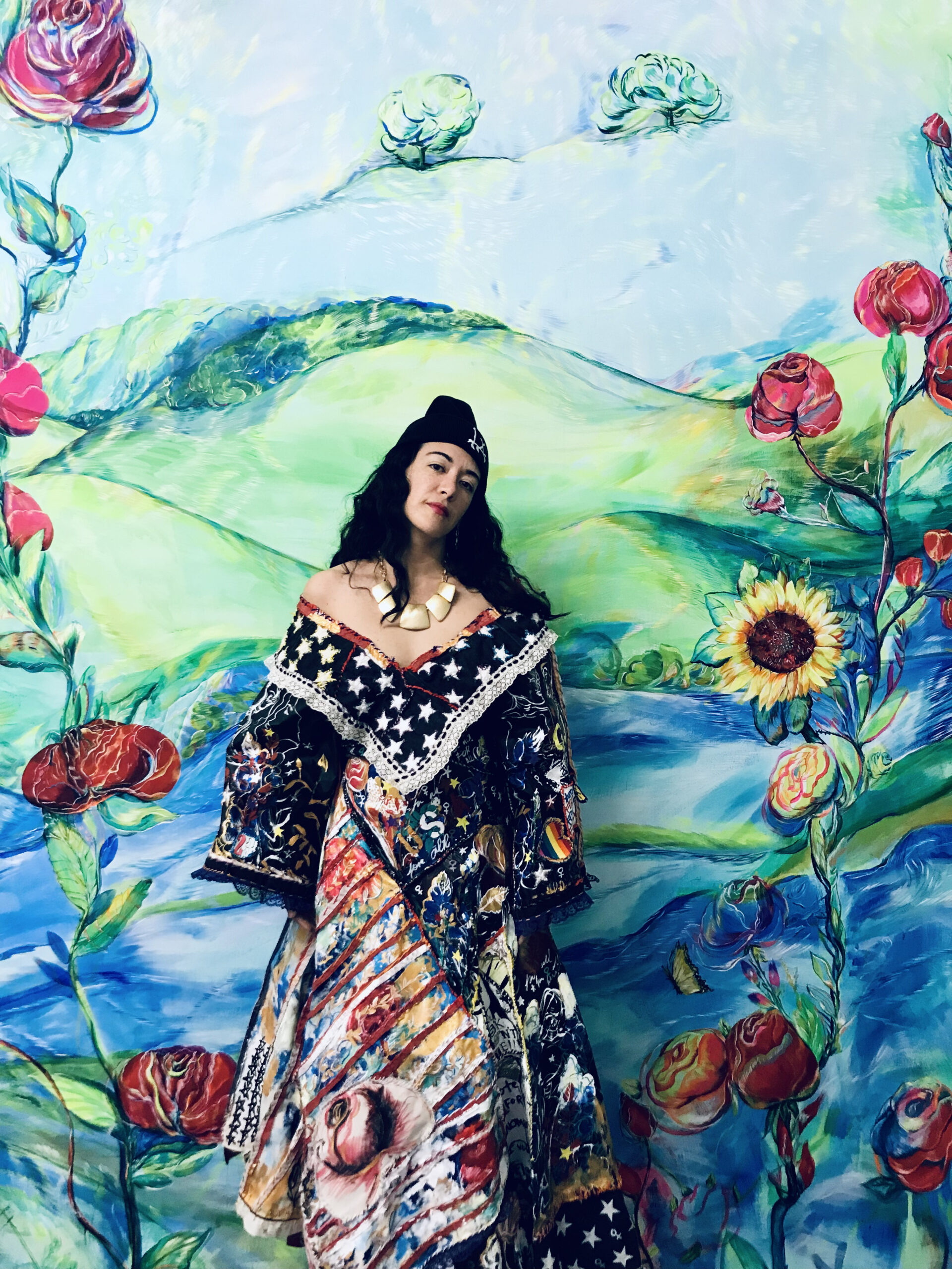 A woman in a beanie, necklace, and dress featuring paintings, stars, and a patchwork of other ephemera, standing in front of a large painting of a rural landscape embellished with roses and sunflowers.