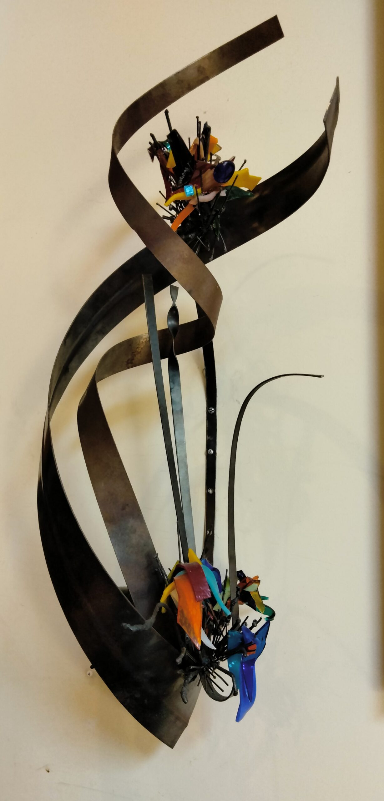 An abstract wall sculpture made up of bended metal and various other colorful materials.