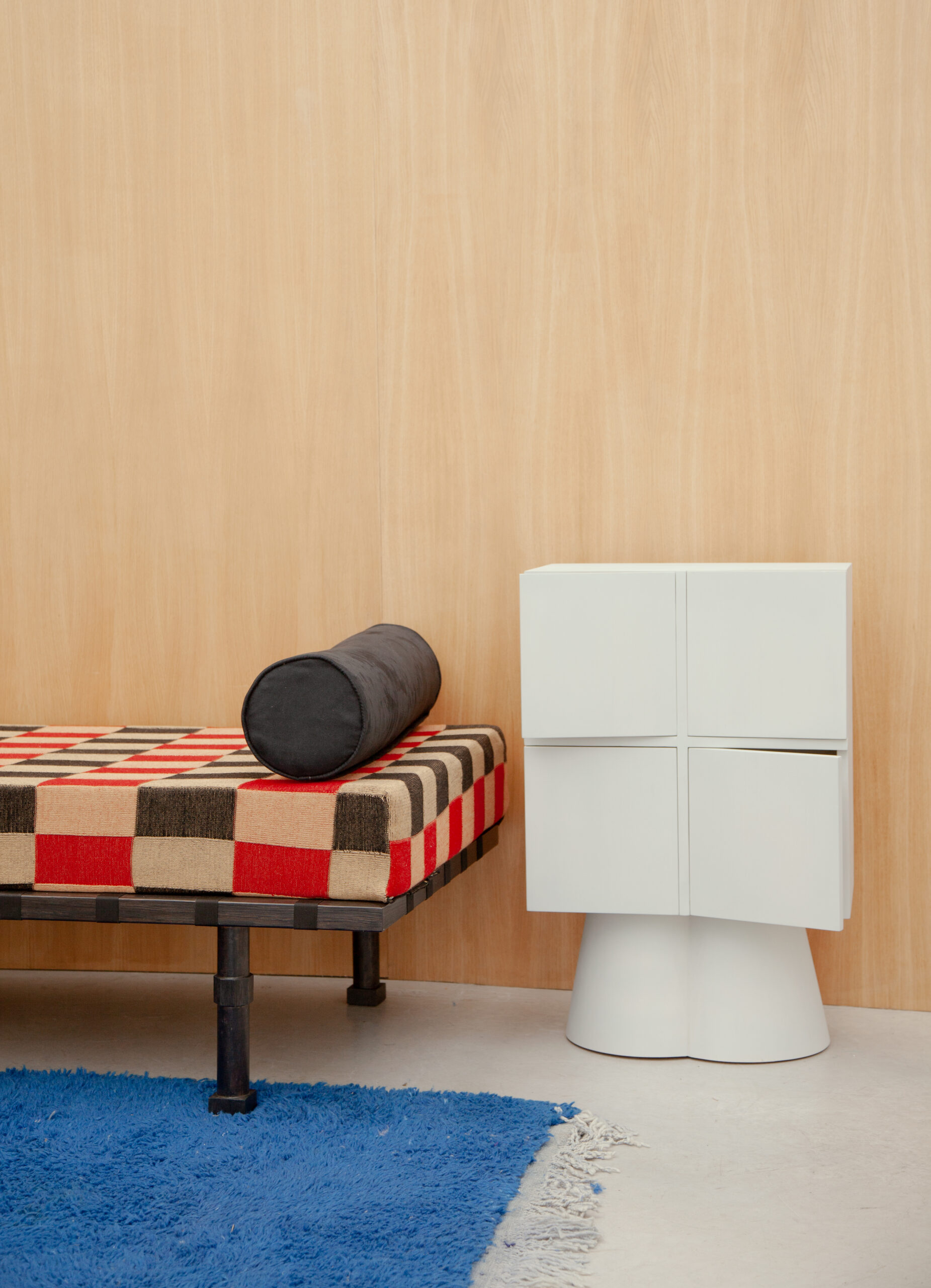 A black and red checkered bed, with a black cylindrical pillow, beside in a square white drawer, in a model room.