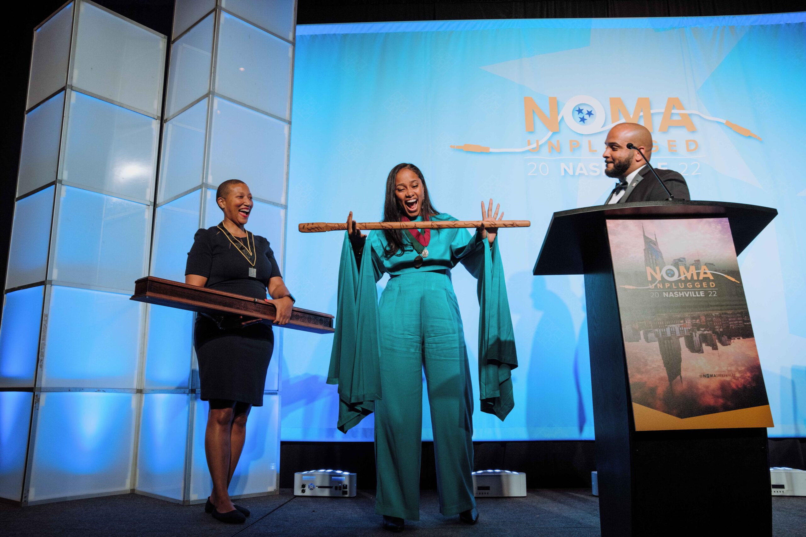 Alumna Pascale Sablan onstage at a ceremony, in an all teal outfit between two people wearing all black, gleefully holding a large wooden staff with engravings on it.