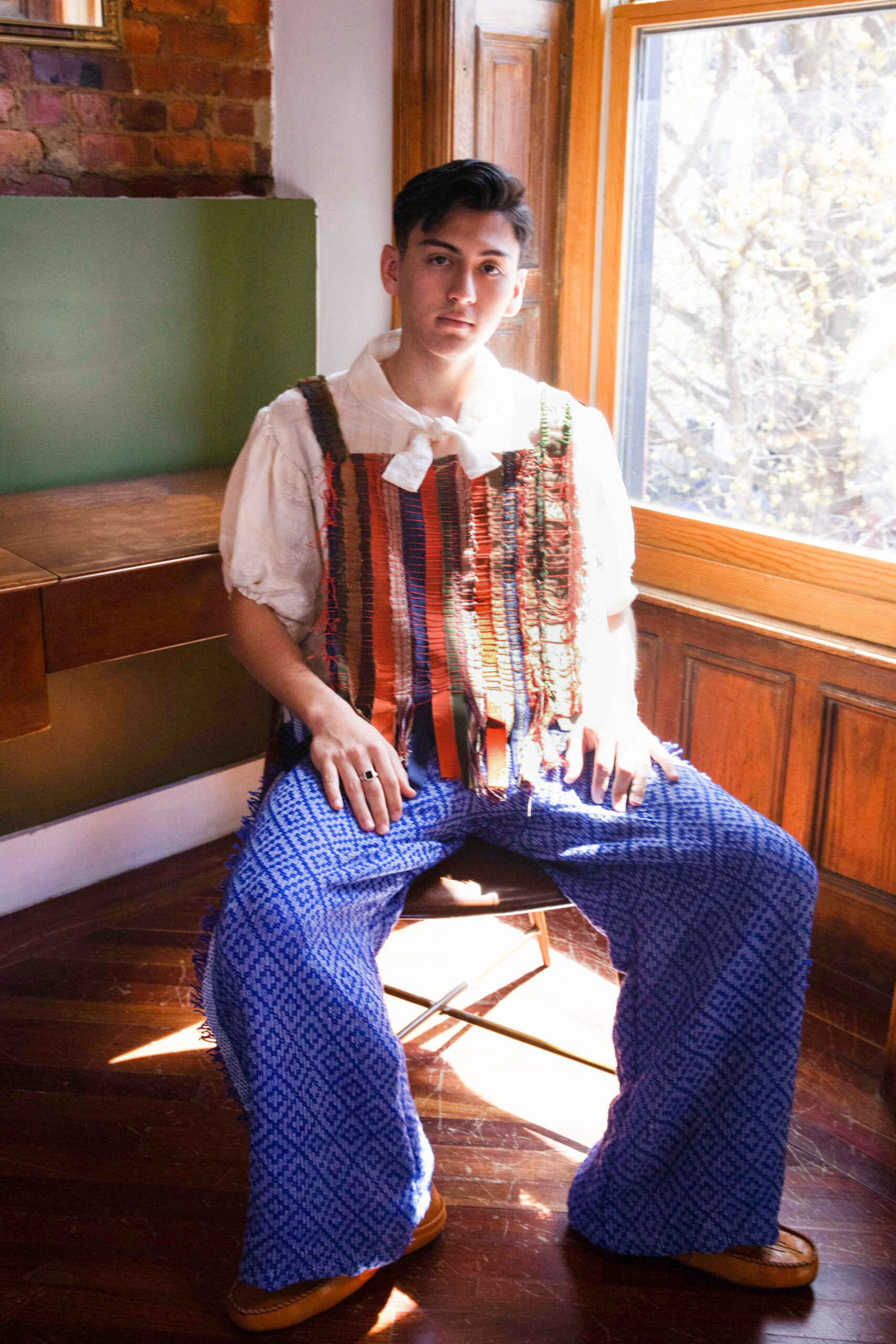 Modeling a look composed of wide blue pants, a white blouse with bow, and a vest made of variously colored ribbons woven in stripes, a model reclines on a chair washed in natural light.