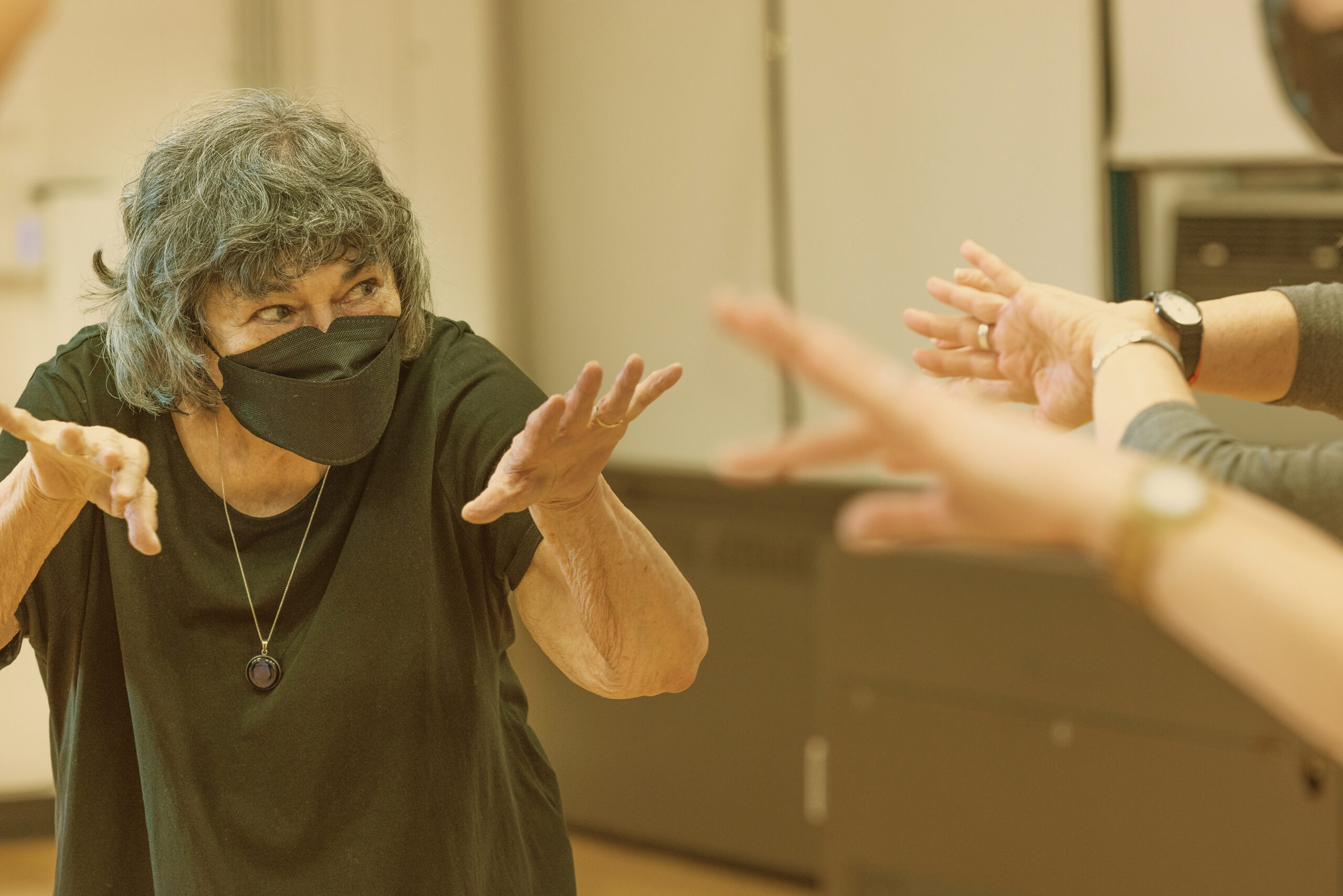 A person in a KN95 mask, black t-shirt, and black necklace crouches with outstretched hands, looking at two people with their hands similarly outstretched but whose bodies are otherwise out of frame.