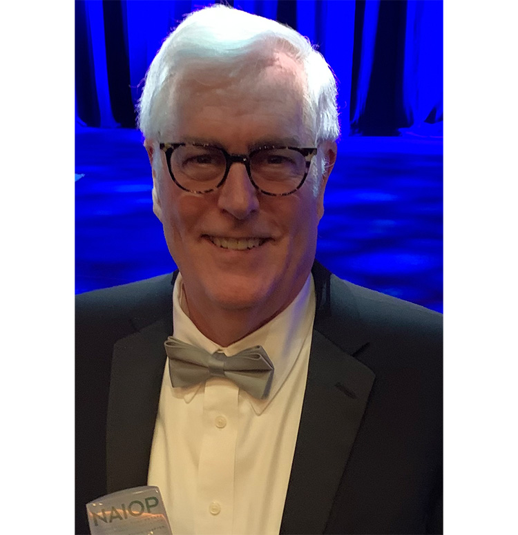 A headshot of a man in a suit with a bowtie and a "NAIOP" name tag, in front of a blue background.