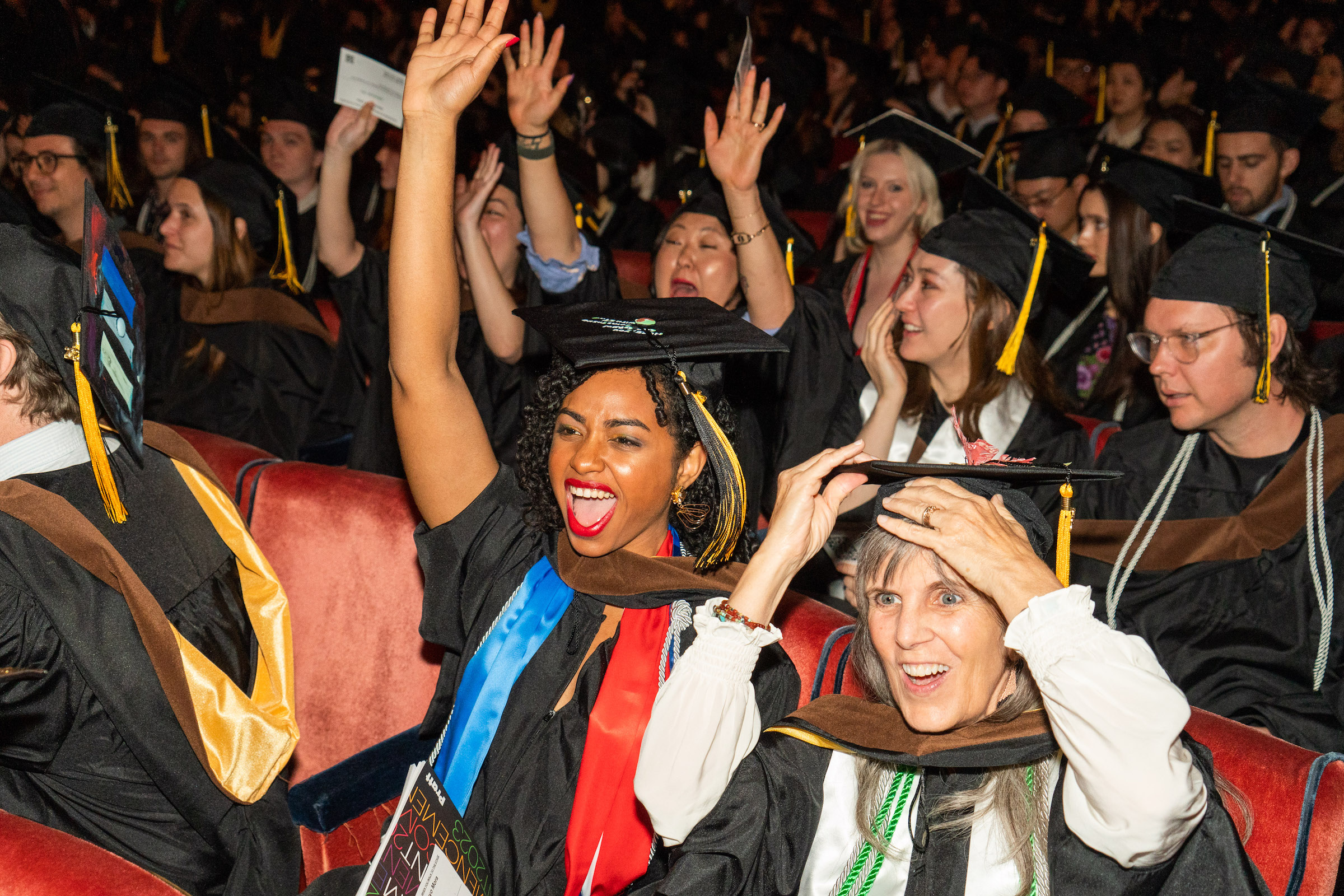 A photograph of graduate in caps and gowns sitting in seats at Radio City Music Hall during the 2023 Commencement ceremony. They are cheering and celebrating with their arms raised in the air.
