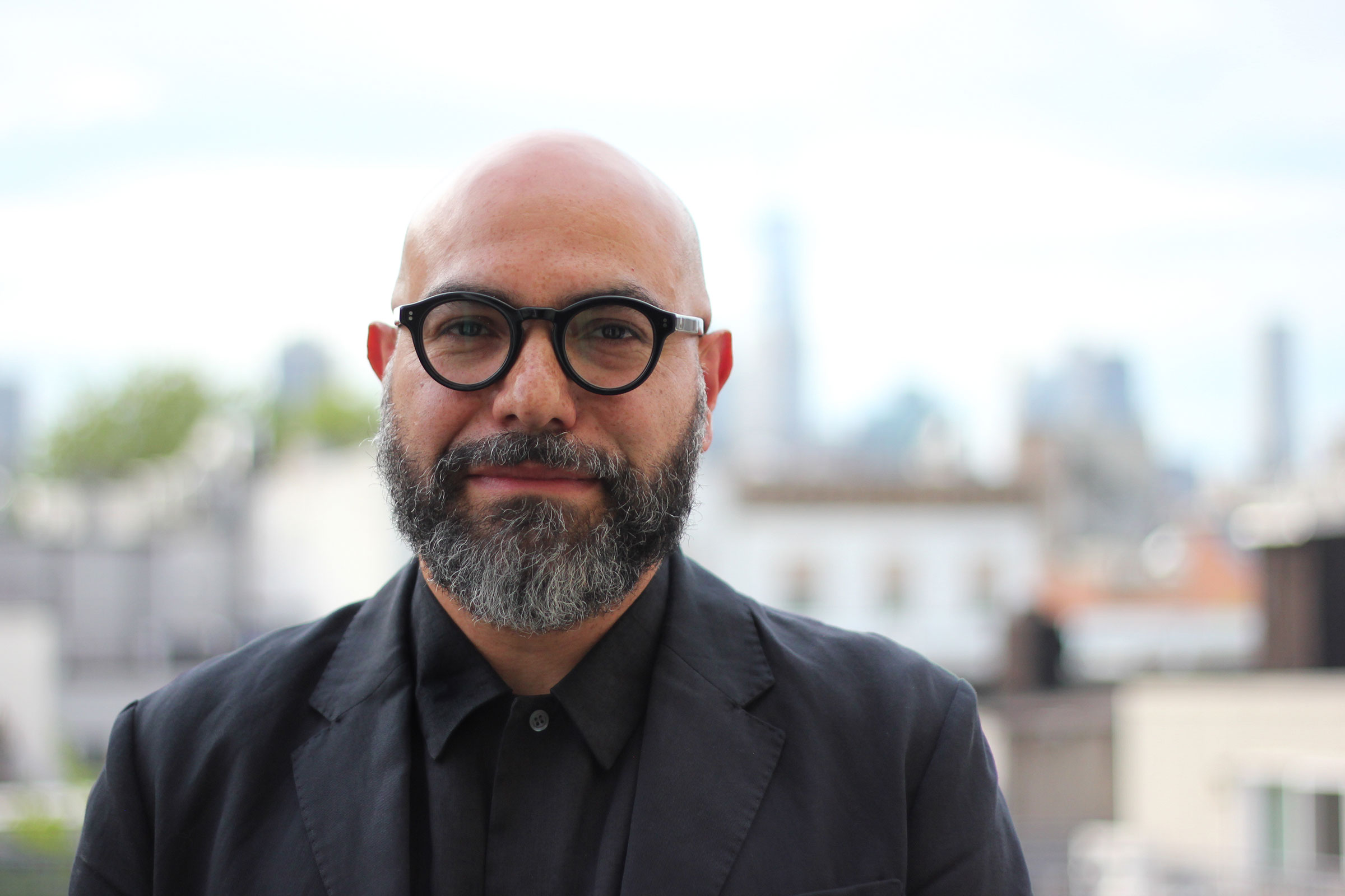 A photograph of Quilian Riano from his chest up. He is looking at the camera, smiling slightly. He's wearing a black jacket over a black shirt that is buttoned up to the top button. Behind him is a blurry skyline of the city.