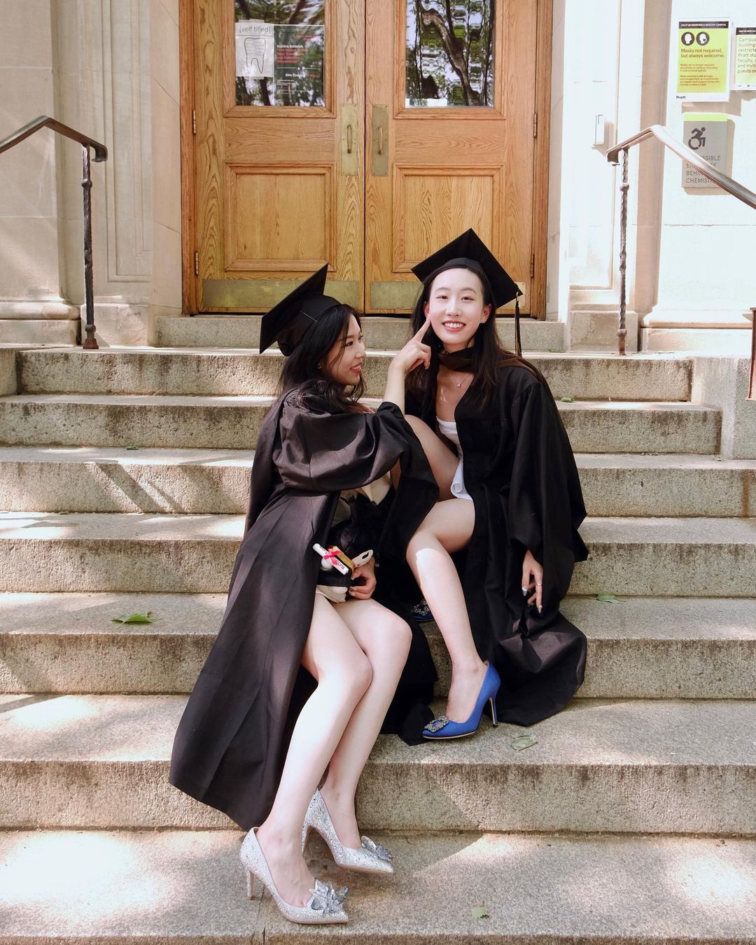 Two students wearing their graduate caps and gowns are sitting on some steps on campus. One is looking at the camera while the other is looking at her friend and pointing at her friend's cheek.