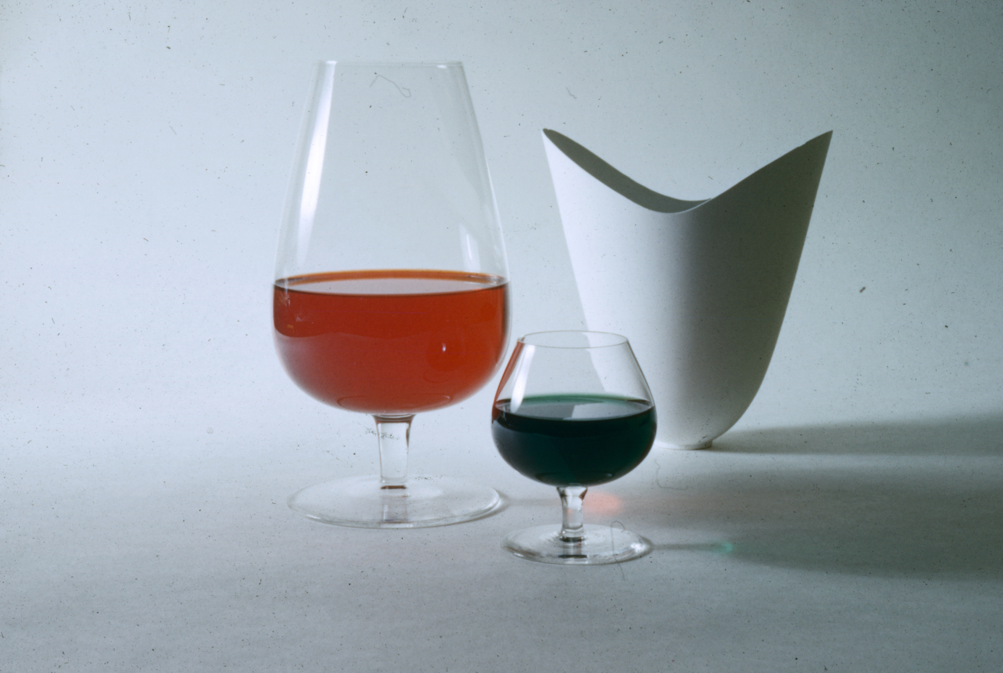 a large glass goblet filled with red liquid, a matching, smaller wine glass filled with dark red liquid, and a white tulip-shaped vase