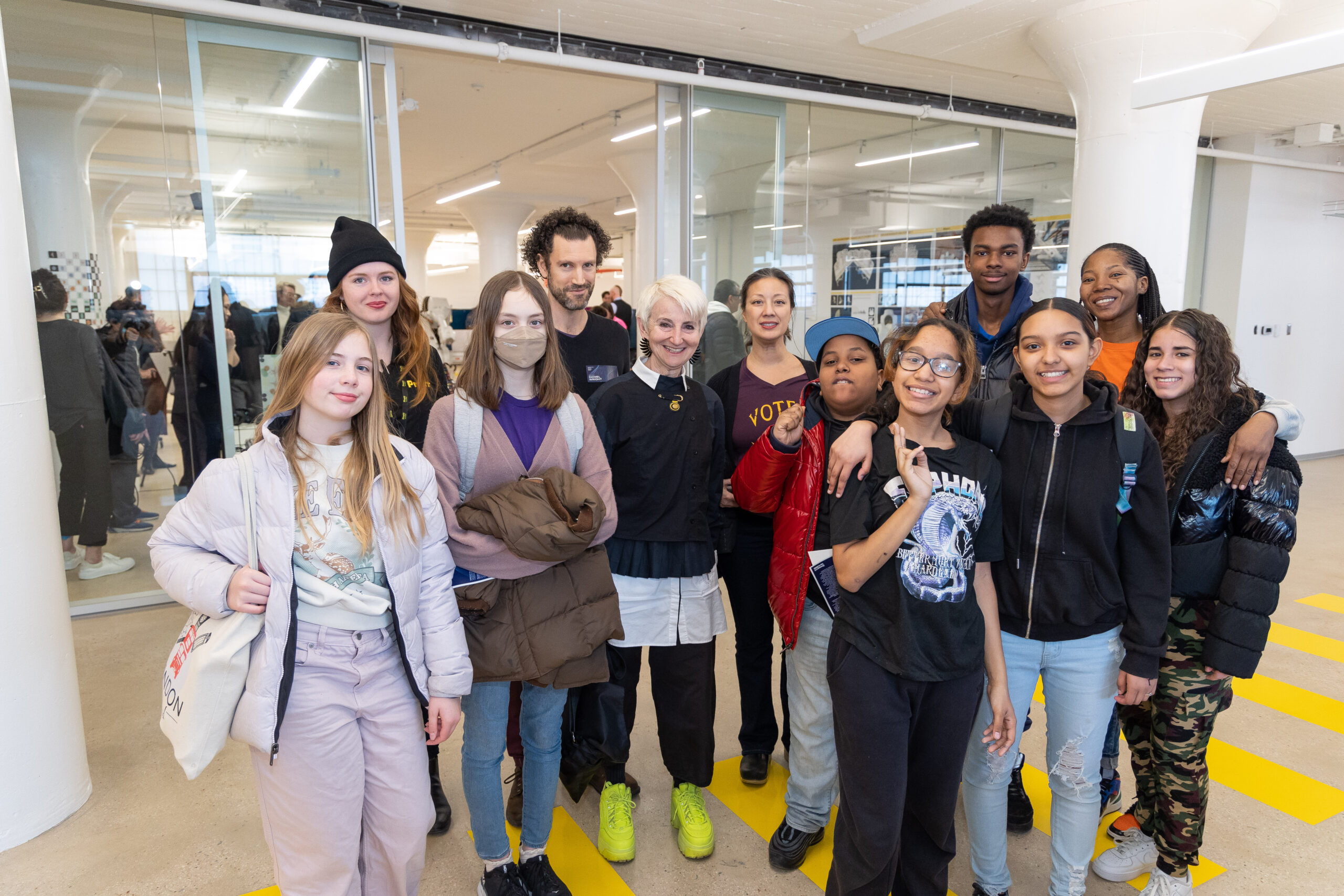 Smiling young people and Pratt faculty and staff pose with Pratt President Frances Bronet in a brightly lit industrial loft space.