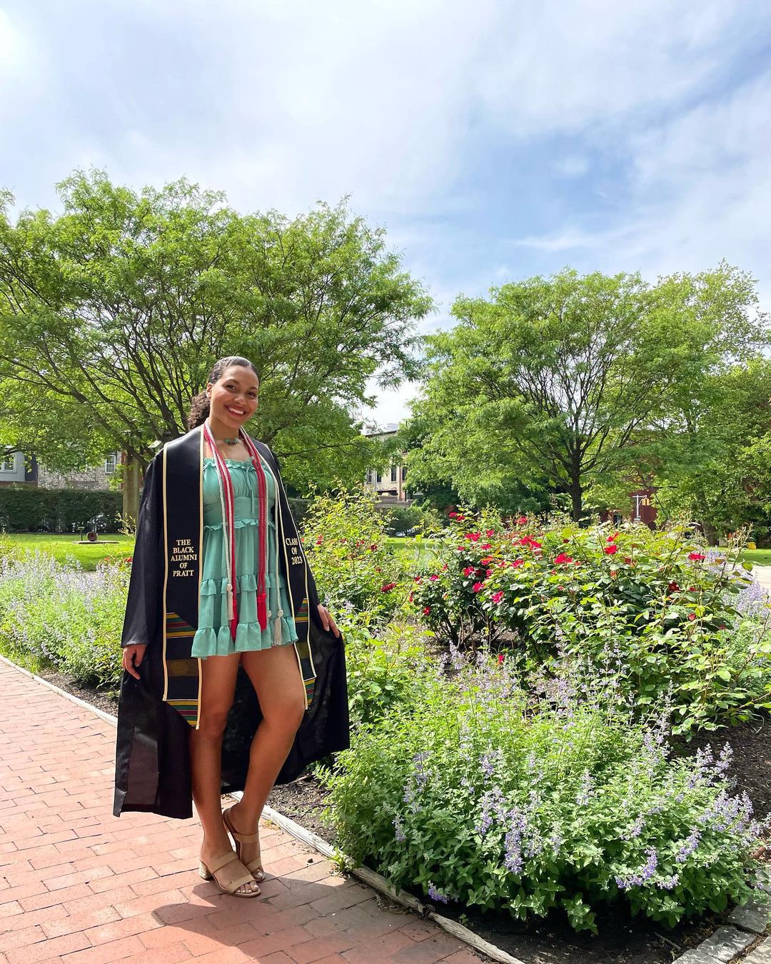 A student in her graduation gown and Black Alumni of Pratt cord stands by the rose garden on campus.