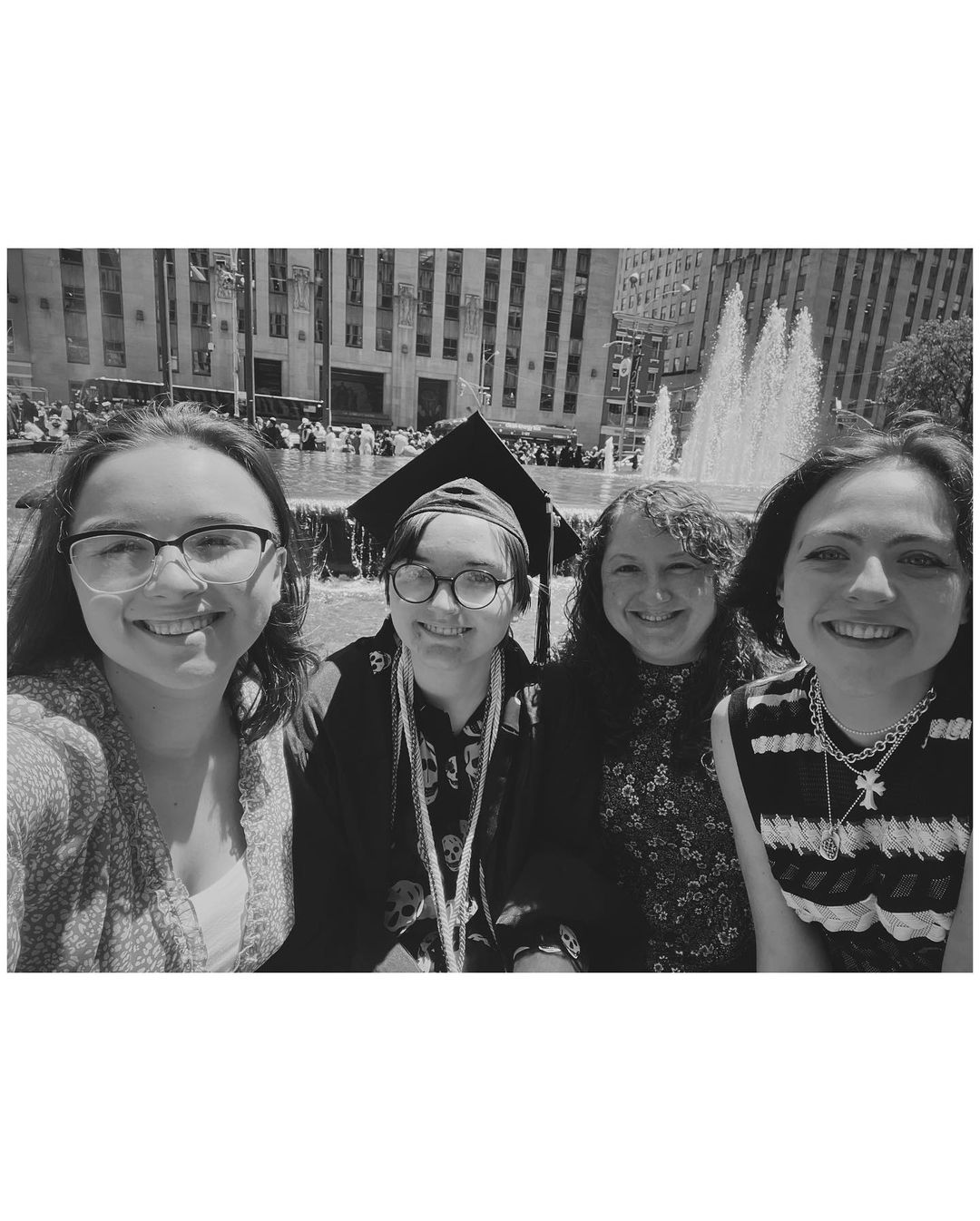 Four students smile at the camera. One is in their cap and gown. They are in front of a fountain in midtown New York City.