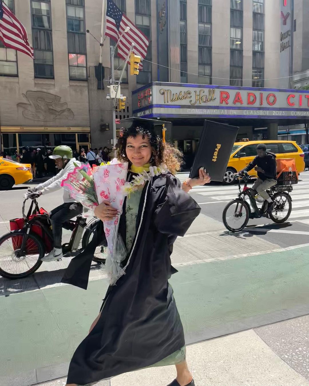 A graduating student in their cap and gown poses in front of the Radio City Music Hall marquee. They are holding flowers and their diploma. Taxis and cyclists are passing her in the street between her and the marquee.