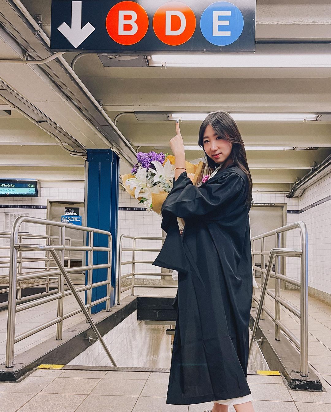 A graduating student in her gown, holding a bouquet of flowers, stands in a subway station beneath a way-finding sign for the B, D, and E trains that also includes a down arrow. She is pointing up at the sign.