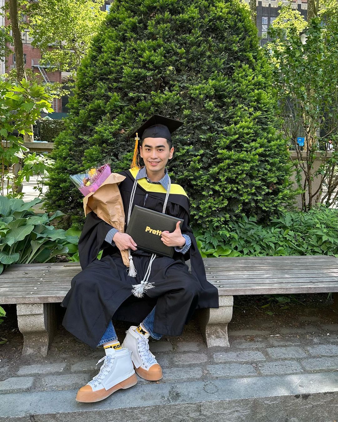 A graduating student dressed in their cap and gown is sitting on a bench surrounded by plants and trees. They are holding a bouquet of flowers and their diploma, smiling at the camera and giving a thumbs up.