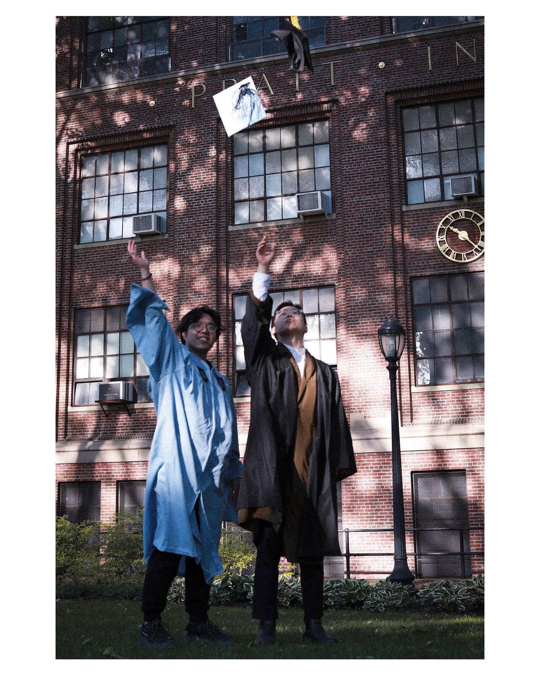 Two students, one dressed in a blue gown, the other in a black one, stand on campus. Their arms are raised as they have just thrown their caps into the air.