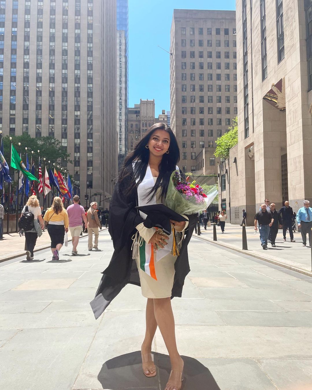 A graduating student stands outside of Rockefeller Center. She is dressed in her graduation gown and is holding a bouquet of flowers and her cap.