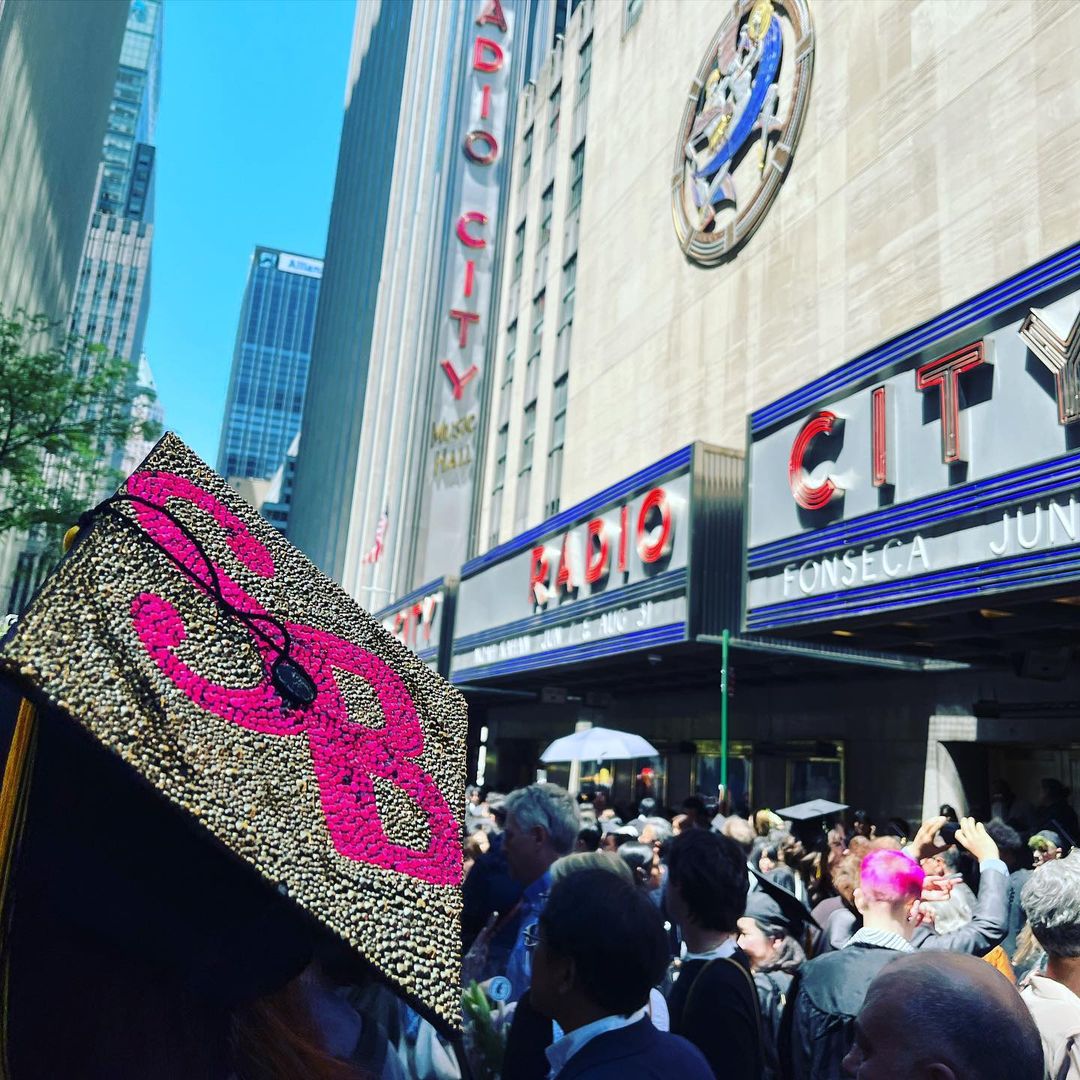 The foreground of the photo shows the top of someone's cap with the letters SB on it. The background shows students standing outside of Radio City Music Hall.