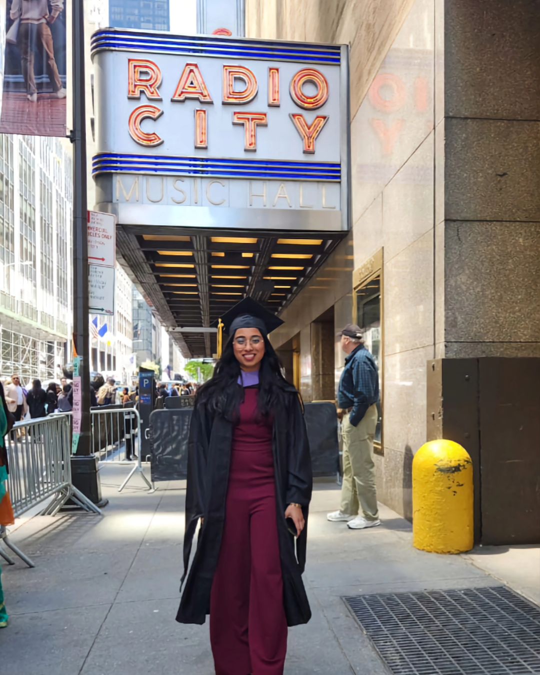 A student dressed in her cap and gown stands in front of the Radio City Music Hall marquee.