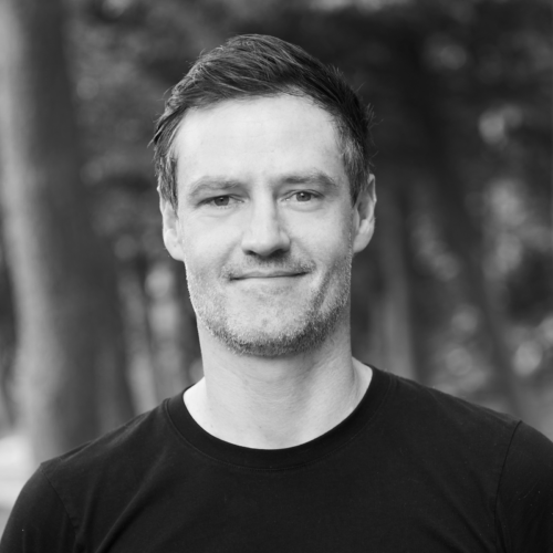 A grayscale portrait of Andre Detller, a man with a close-cropped haircut and beard, wearing a dark turtleneck, looking to the side in a forest setting.
