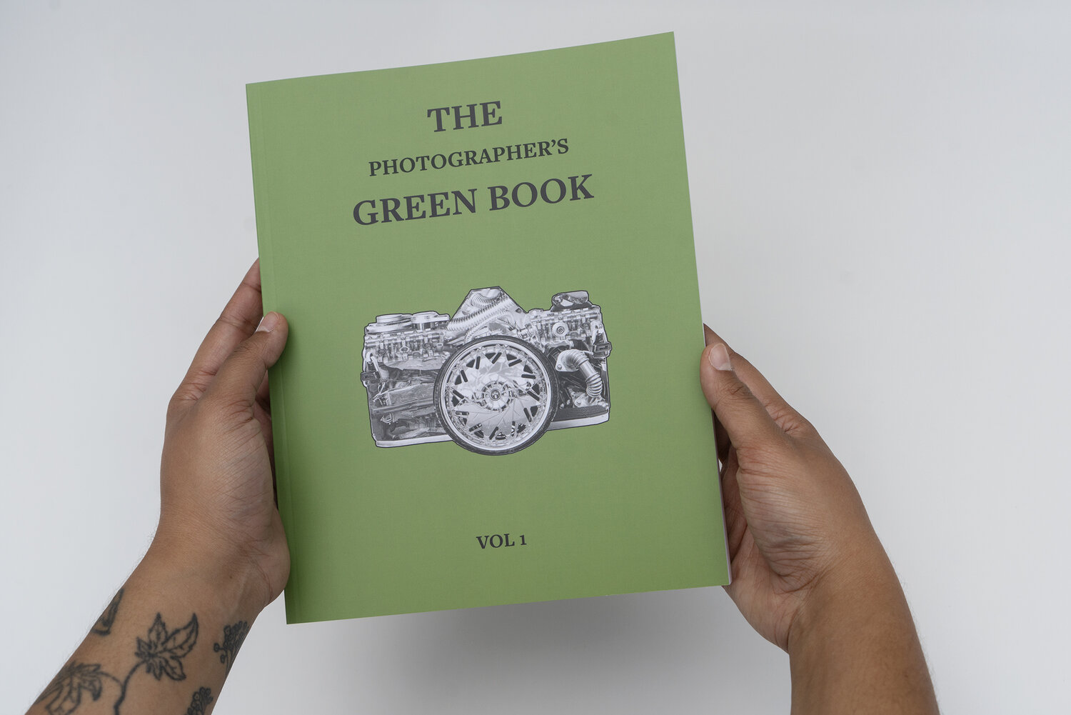 A green book cover with a black and white sketch of a camera containing various patterns on its surface being held by a tattooed person.
