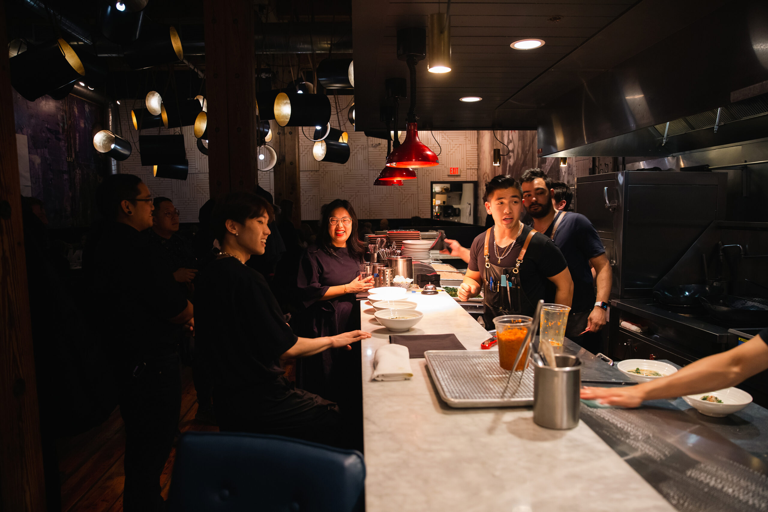 Laughing people gather around the bar of a restaurant's open kitchen