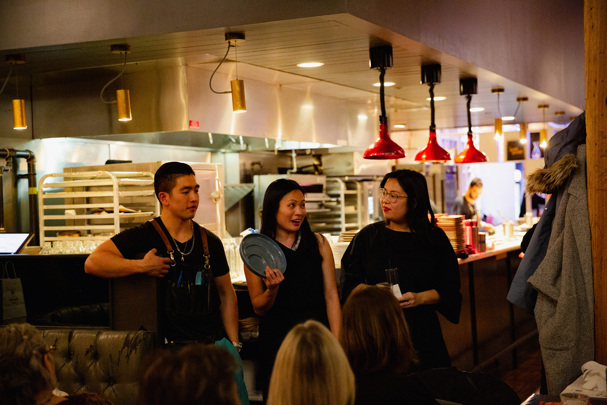 Two designers and a chef stand before open kitchen in a warmly lit restaurant, speaking to an audience of diners