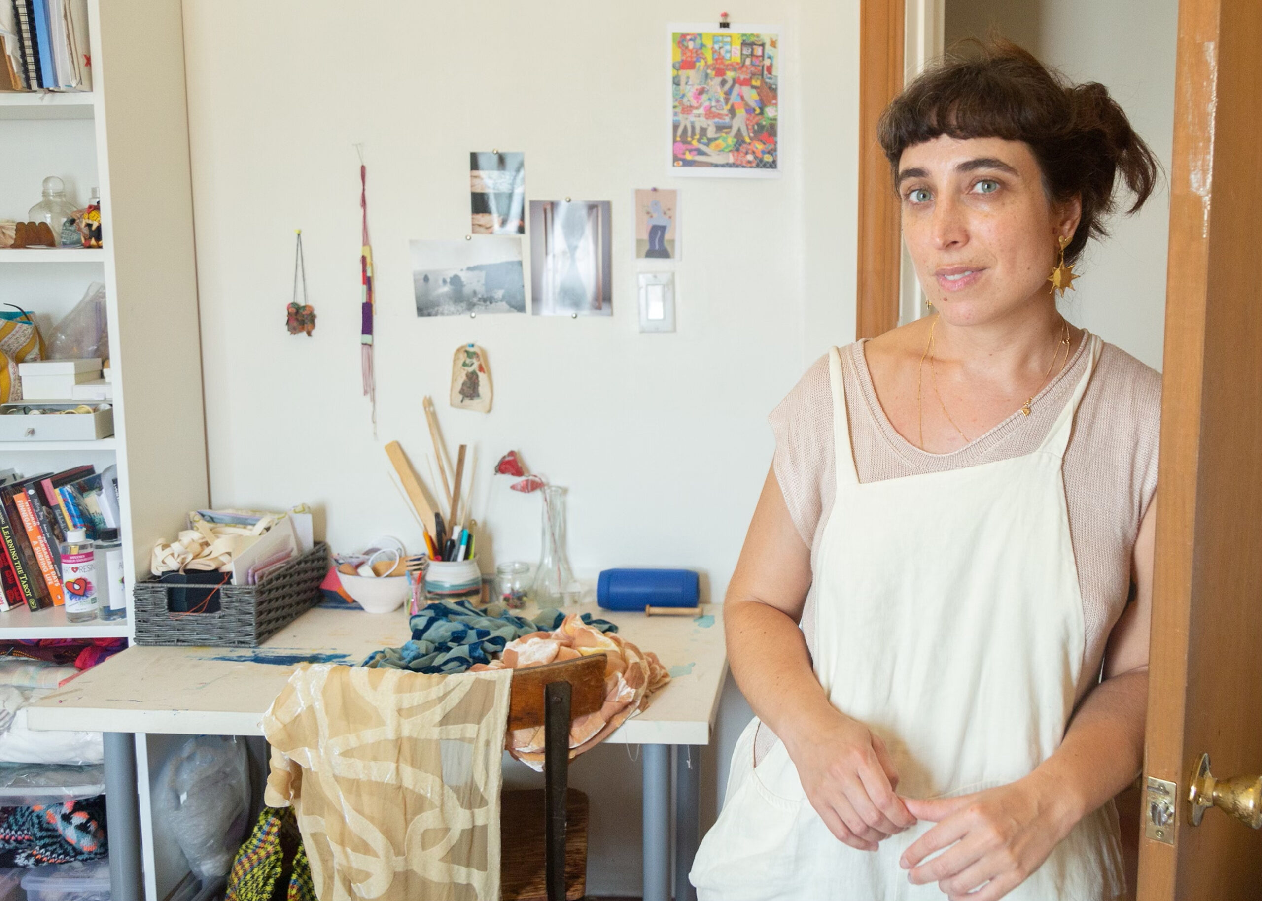 Isa Rodrigues stands in front of a work table with fabrics and tools, facing a wall with reference imagery and objects