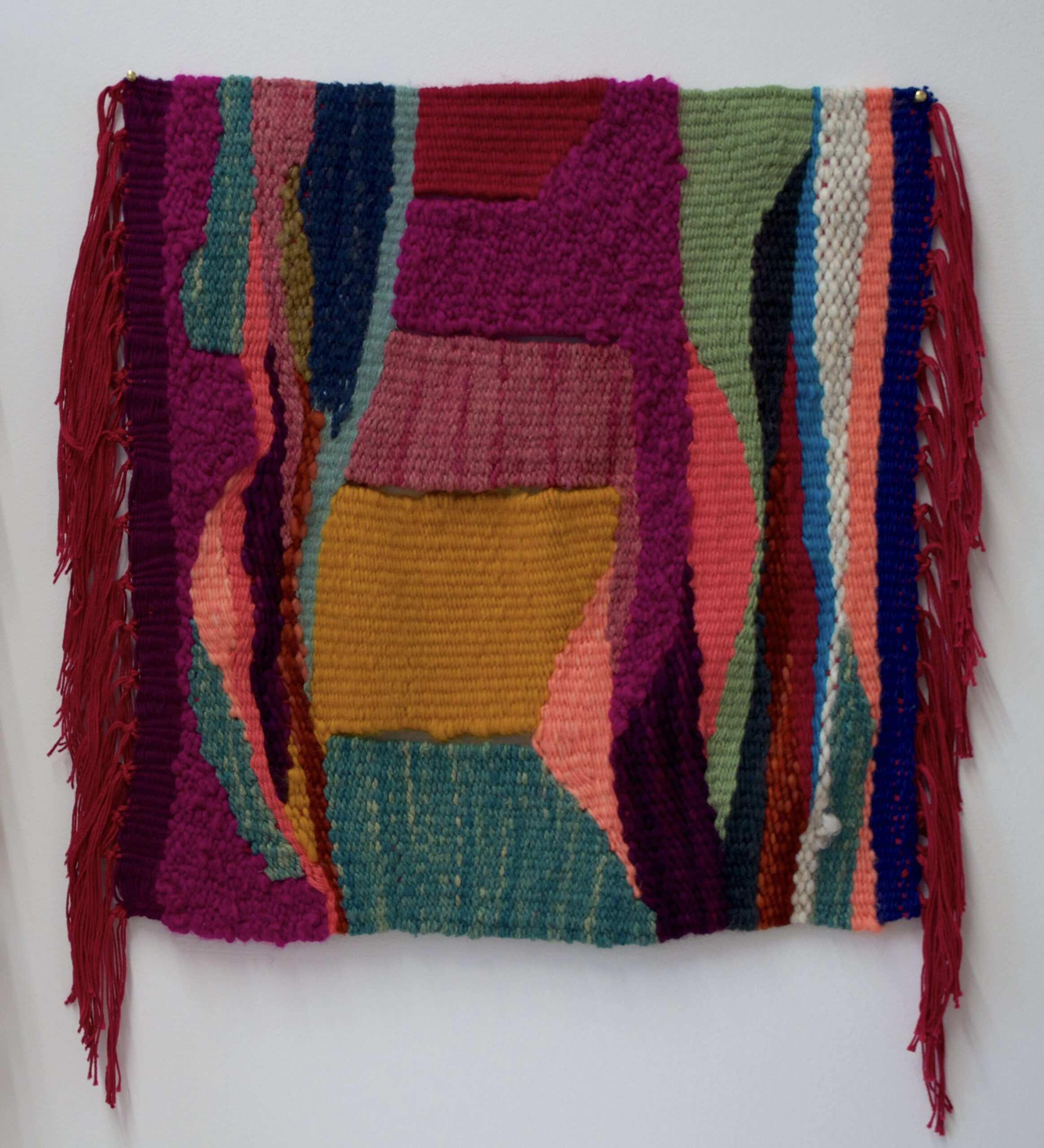 Woven tapestry with deep red, green, and black fibers forming abstract shapes interspersed with slivers of brighter coral, blue, and moss