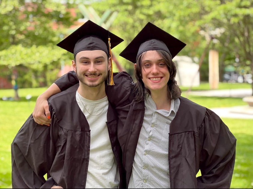 Two graduating students wearing their caps and gowns are standing together on campus smily at the camera. One has their arm around the other's shoulders.