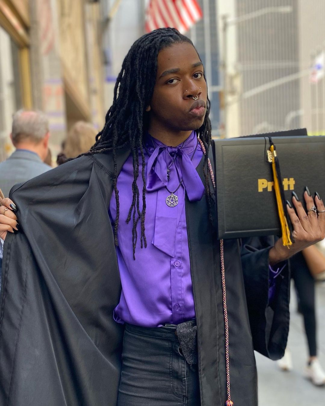 A graduating student is wearing their gown and looking at the camera. They are opening the front of their gown to show off their outfit: a purple shirt and necklace depicting a pentagram. They are holding their diploma in their other hand.