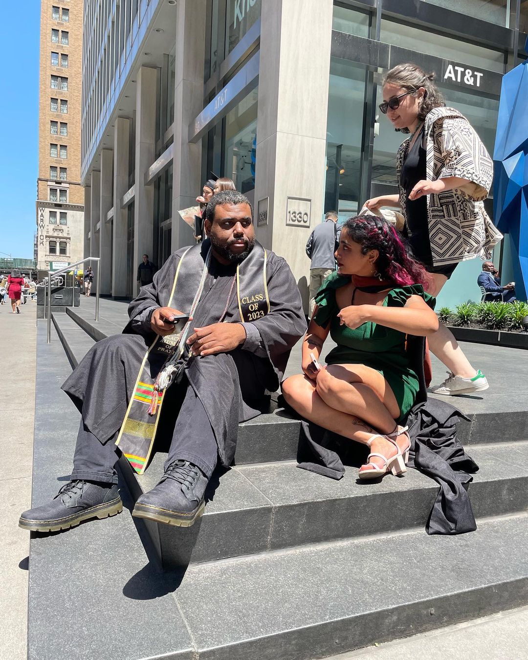 A student wearing their graduation gown and Black Alumni of Pratt cord is sitting on some steps in midtown Manhattan. A friend is sitting next to them and they are having a conversation. Another friend is walking around behind them.