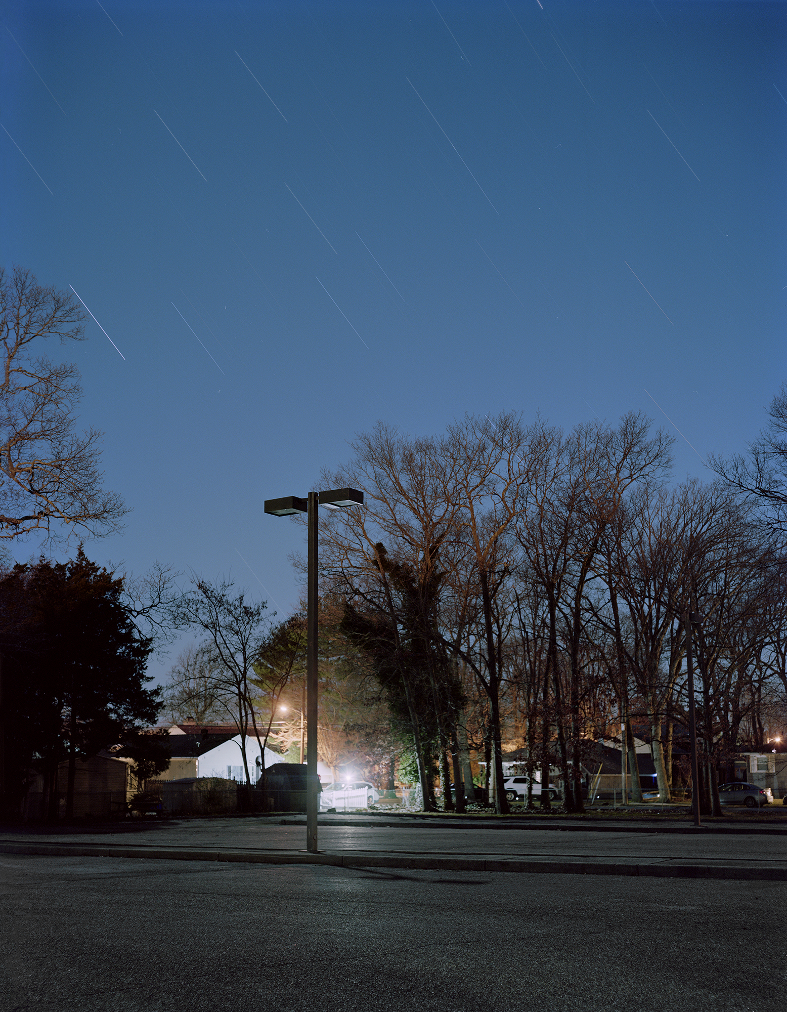A long exposure of a parking lot with trees behind it. A lone streetlight is in the center of the parking lot. The streaks of a stars in the sky can be seen.