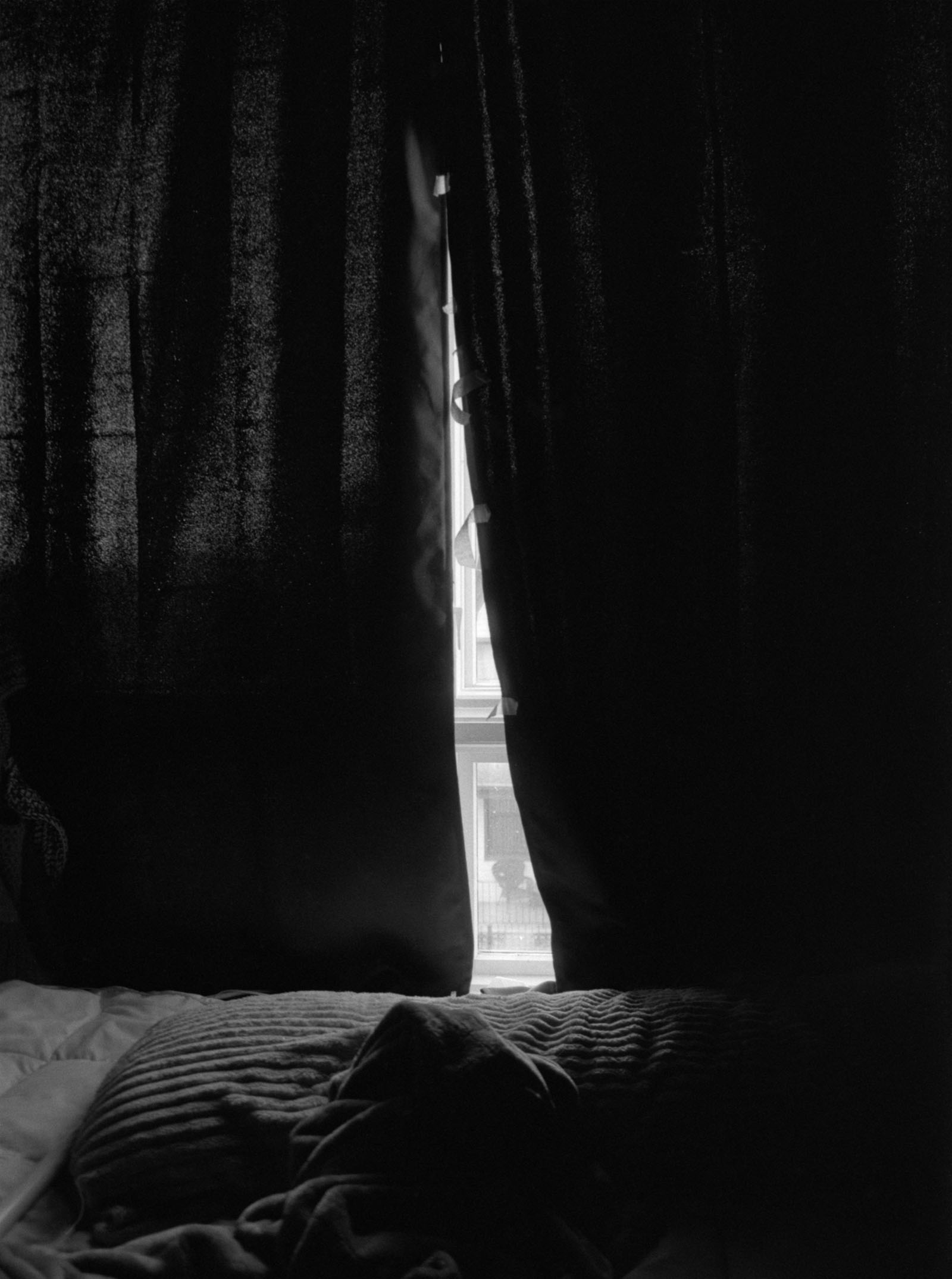 A black and white photograph showing a bed and some curtains covering a window behind it. There is a sliver of light coming through the center of curtains.