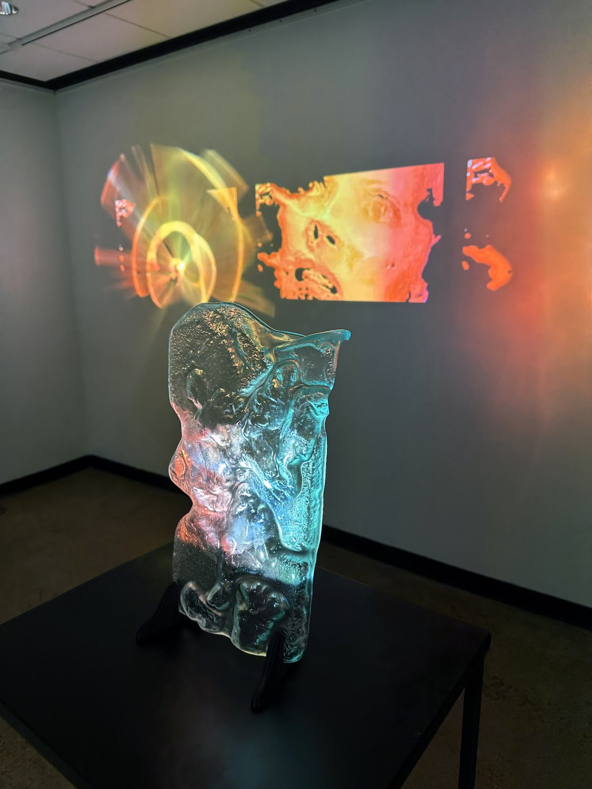 projection of womans face, with post-processing added on wall, behind a clear sculpture