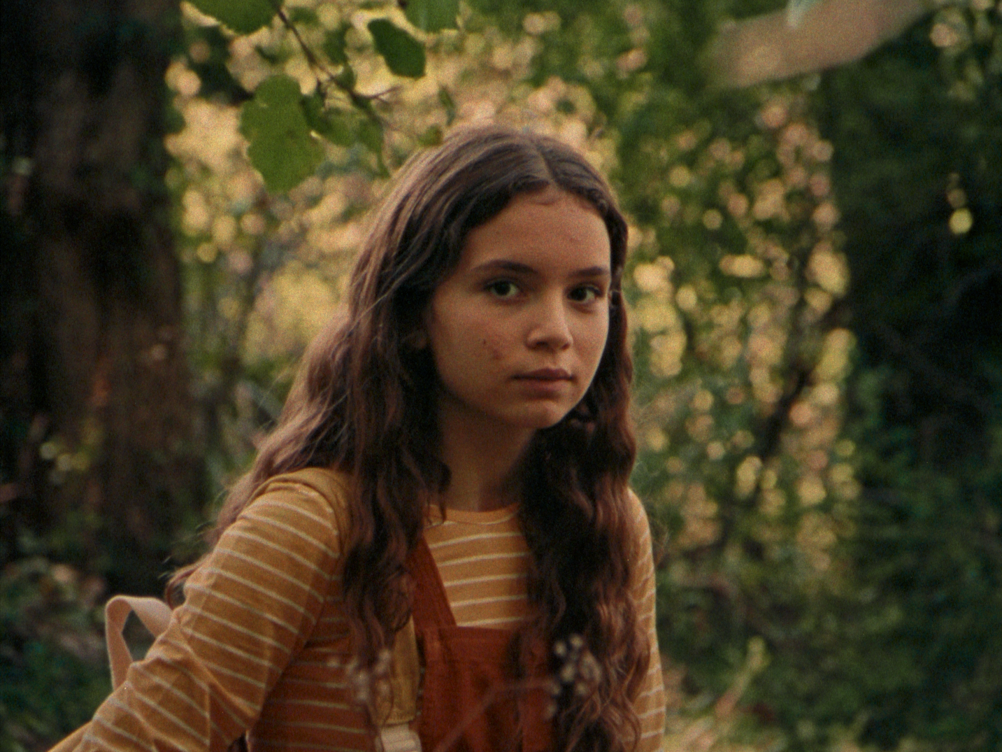 A girl sits in a forest while looking at the camera with a despondent face
