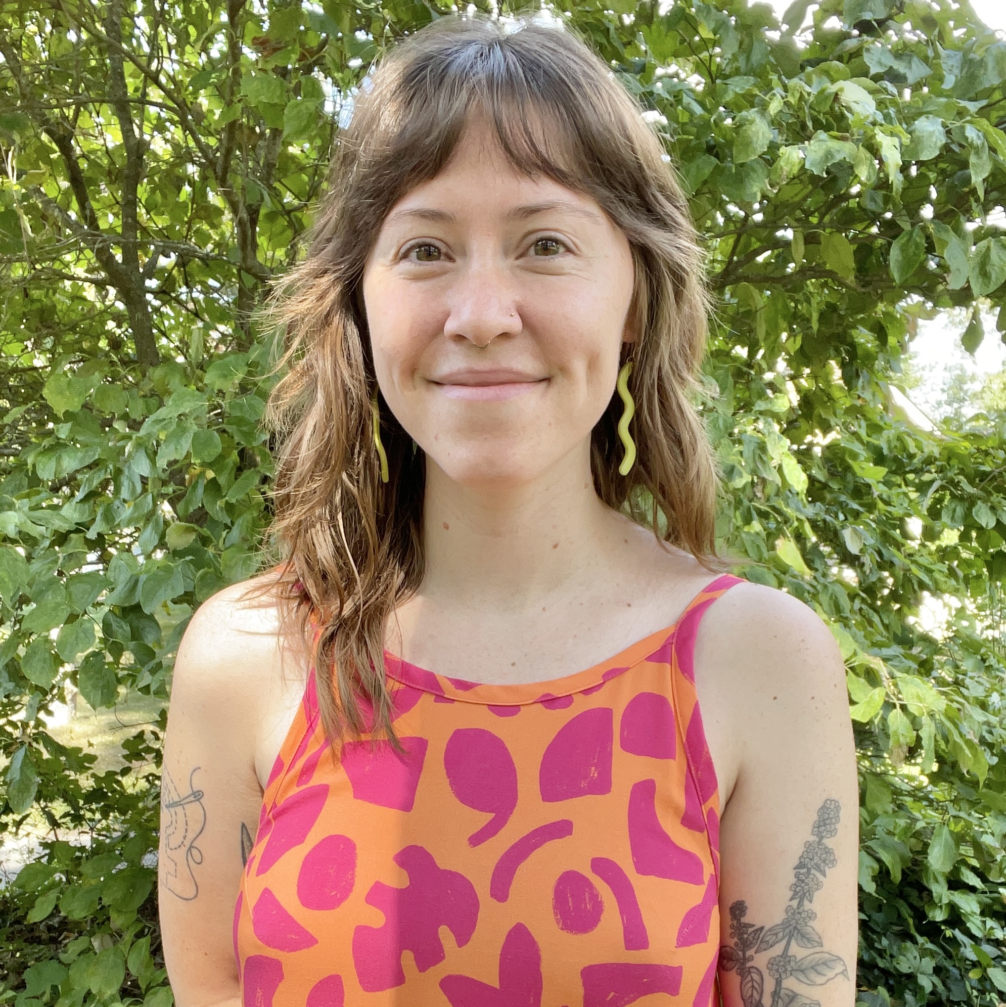 headshot of Gabriella Evergreen, standing in front of lush foliage, wearing a multi-colored top