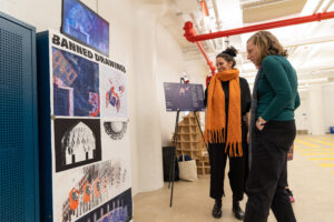two people discuss artwork they are looking at, in a gallery room, one woman wears large scarf and all black clothing, other woman wearing large coat and autumn winter clothing