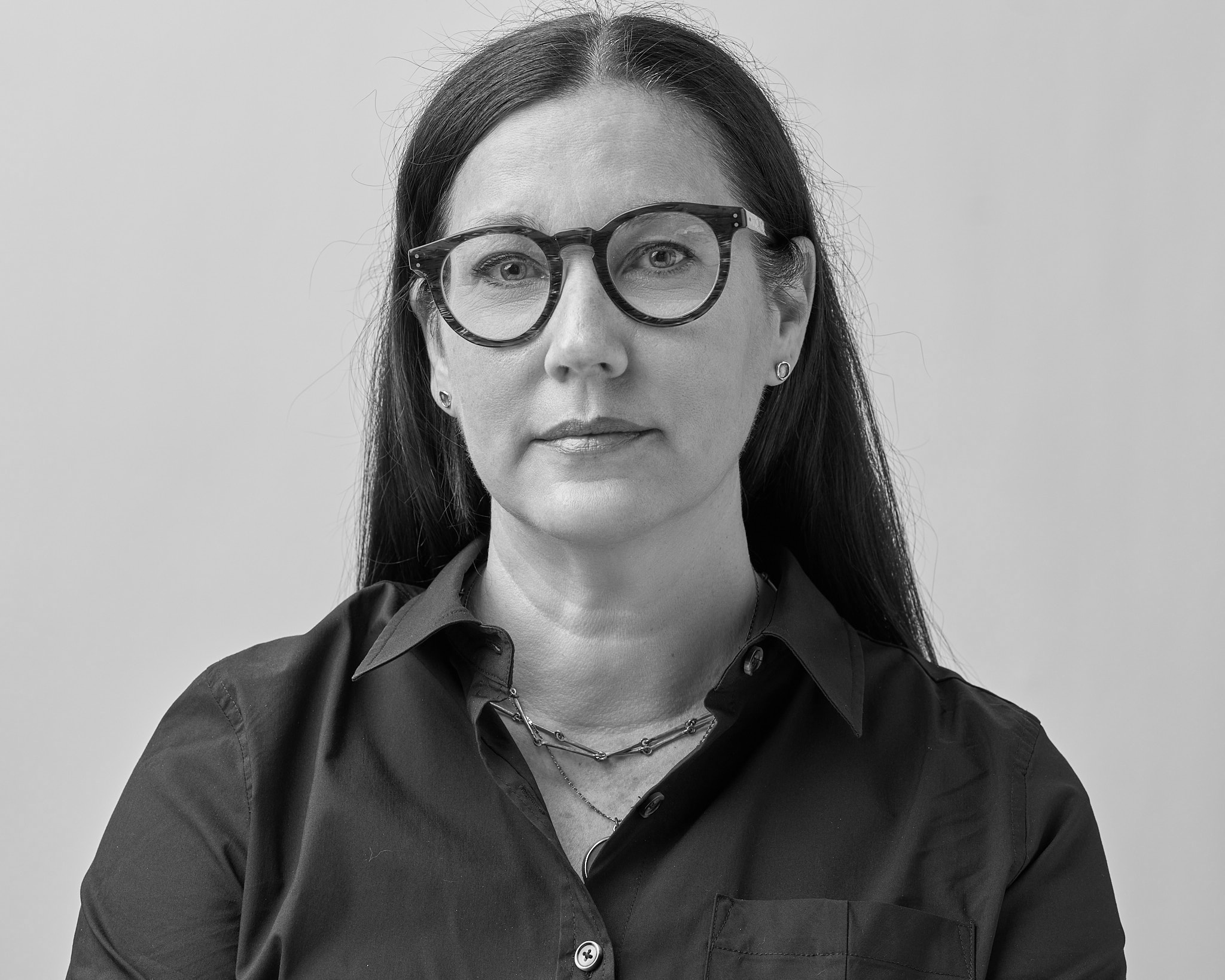 Black-and-white studio portrait of a woman with long straight hair wearing round glasses and a dark button-up shirt, looking directly at the camera against a plain light background.