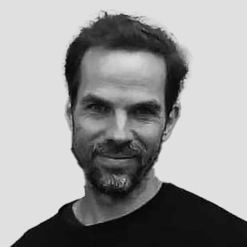 A black-and-white portrait of a man with a short beard and mustache, wearing a black shirt. He has a friendly smile and looks directly at the camera. The background is a plain, light gray color.