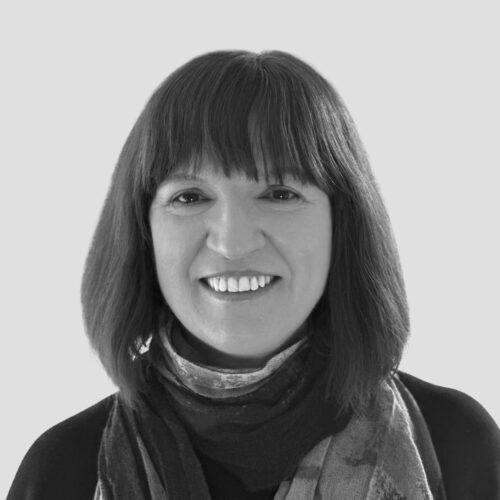A black-and-white portrait of a woman with shoulder-length dark hair and bangs. She is wearing a dark top with a patterned scarf wrapped around her neck. She has a warm smile and looks directly at the camera. The background is a plain, light gray color.