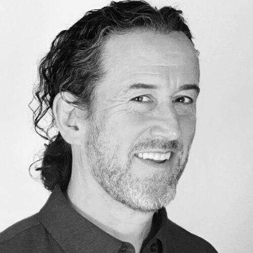 A man with shoulder-length curly hair and a salt-and-pepper beard smiles while looking slightly off-camera. He wears a dark collared shirt and stands against a bright, plain background. The photo is in black and white with soft contrast.