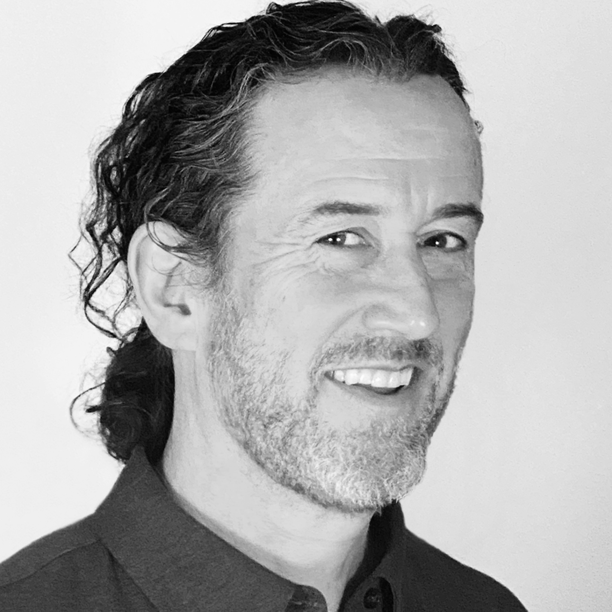 A man with shoulder-length curly hair and a salt-and-pepper beard smiles while looking slightly off-camera. He wears a dark collared shirt and stands against a bright, plain background. The photo is in black and white with soft contrast.