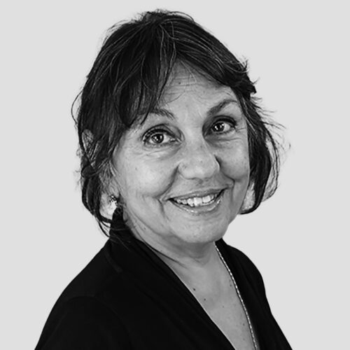 A black-and-white portrait of a woman with shoulder-length, wavy dark hair. She is wearing a dark top with a necklace and has a warm smile. The background is a plain, light gray color.