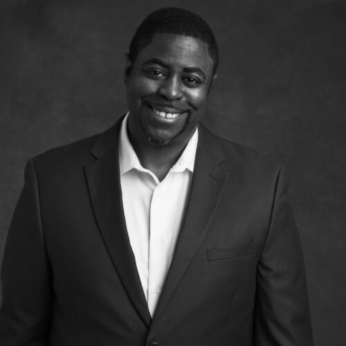 A black-and-white portrait of a man smiling warmly, wearing a dark suit jacket over a light-colored button-down shirt. He is posed against a softly lit studio backdrop, slightly angled to one side with a relaxed and confident demeanor. The lighting highlights his face and upper body, creating a professional yet approachable atmosphere.
