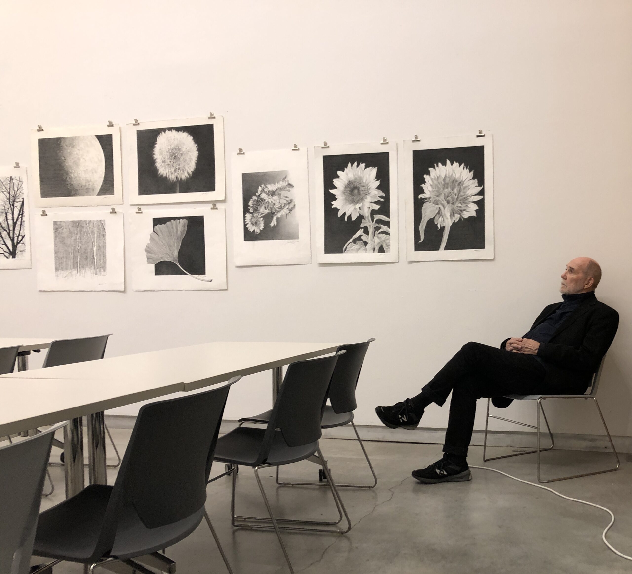 Woody Rainey sits on a char inside a classroom as he looks off to the left. There are drawings and sketches hung on a white wall next to him.