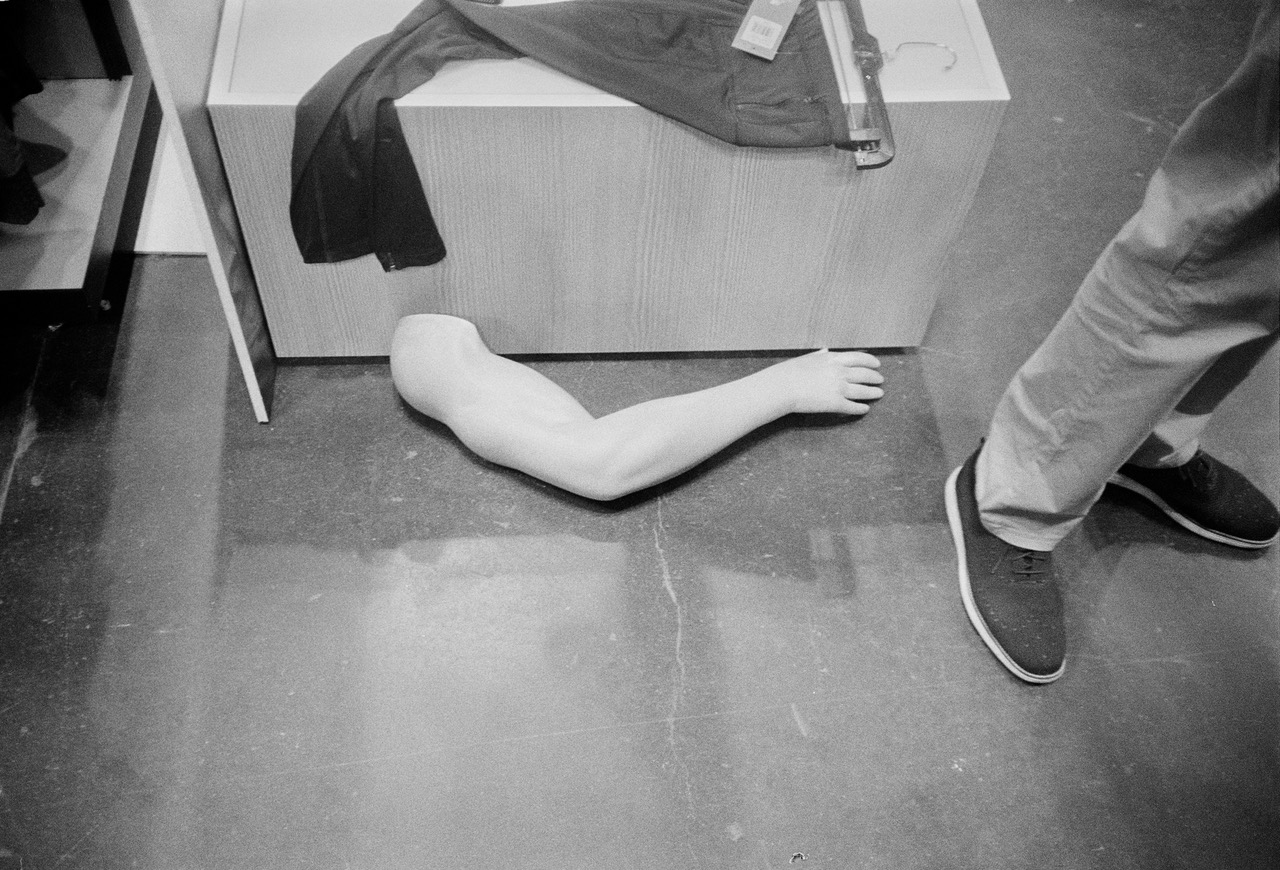 The image shows a black and white image of a mannequin's right arm set down on the floor beside a clothing rack. A man is standing beside the mannequin arm to the right of the image. Only his pant and shoes are visible in the image