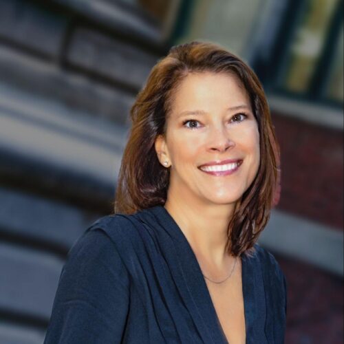 Professional headshot of a woman smiling outdoors, wearing a dark navy dress with a pleated neckline. The background is softly blurred, showing a brick building and architectural details.
