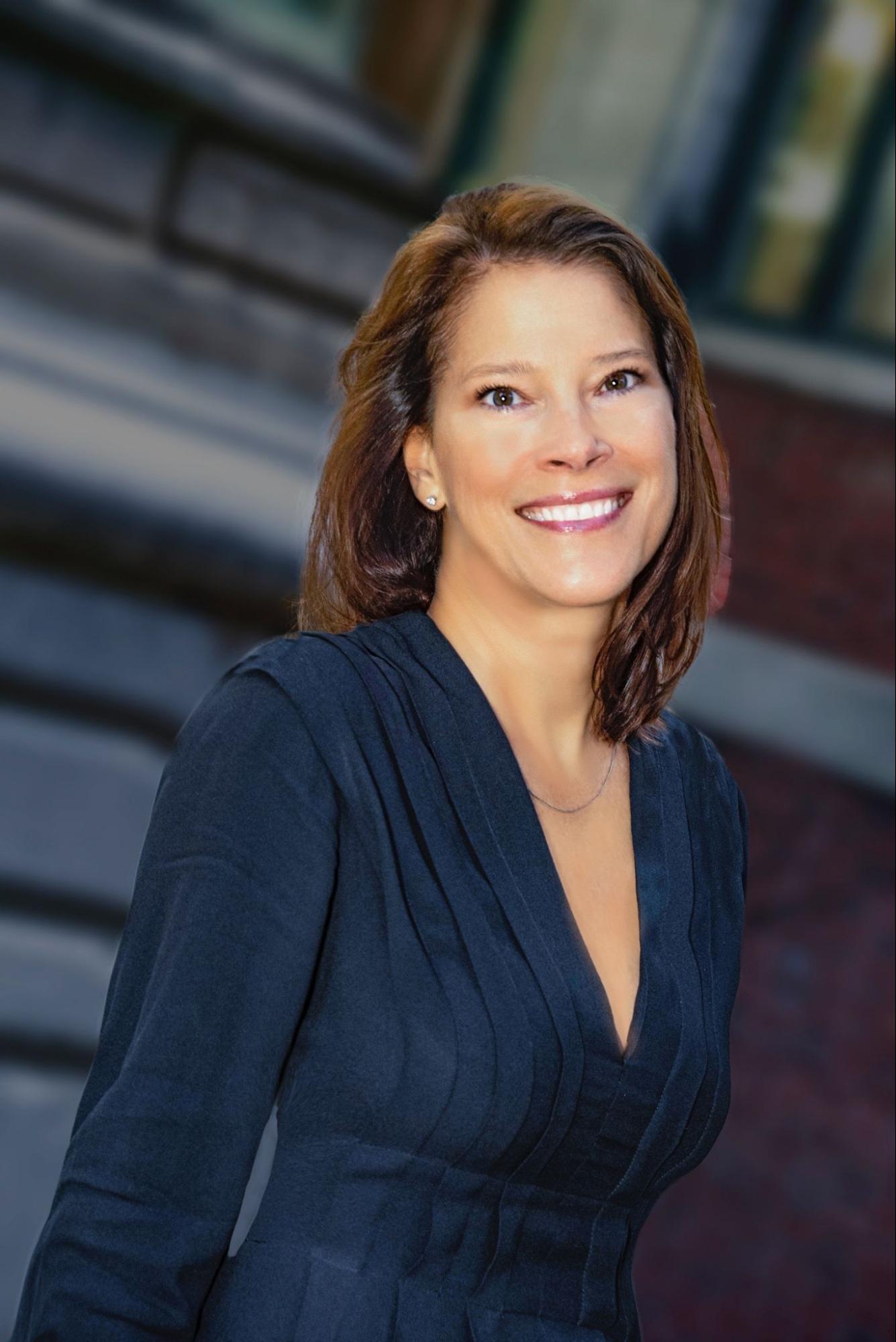 Professional headshot of a woman smiling outdoors, wearing a dark navy dress with a pleated neckline. The background is softly blurred, showing a brick building and architectural details.