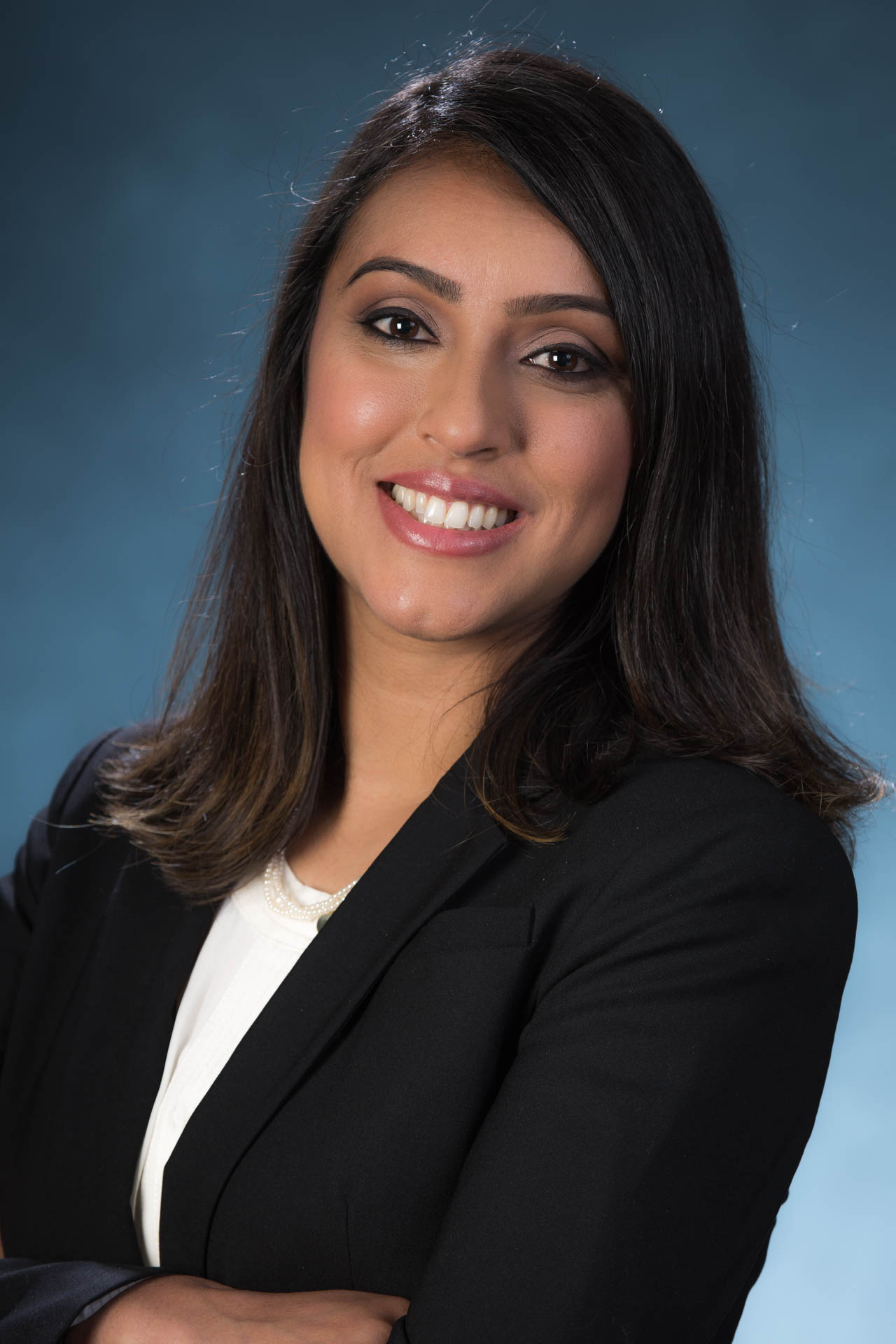Professional headshot of a woman smiling confidently, wearing a black blazer and white blouse against a blue gradient studio background.