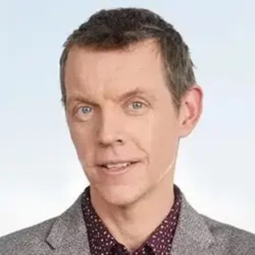 Headshot of a man with short brown hair wearing a gray jacket and patterned shirt, posing against a light background.